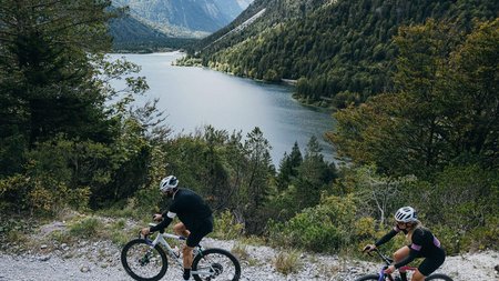 Epic Route Kärnten – Slowenien © WRTG Fotograf Sam Strauss Zwei Radfahrer fahren auf einem Bergweg neben einem See