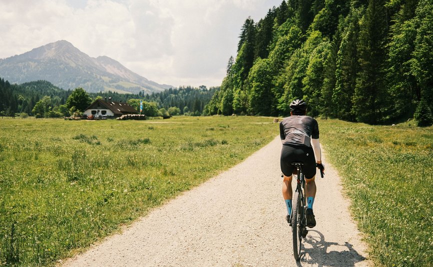 Gravelbike Urlaub in Gravel.Tirol © Liz Kellerer Fahrradfahrer auf einem Weg durch eine grüne Wiese mit Bergen im Hintergrund