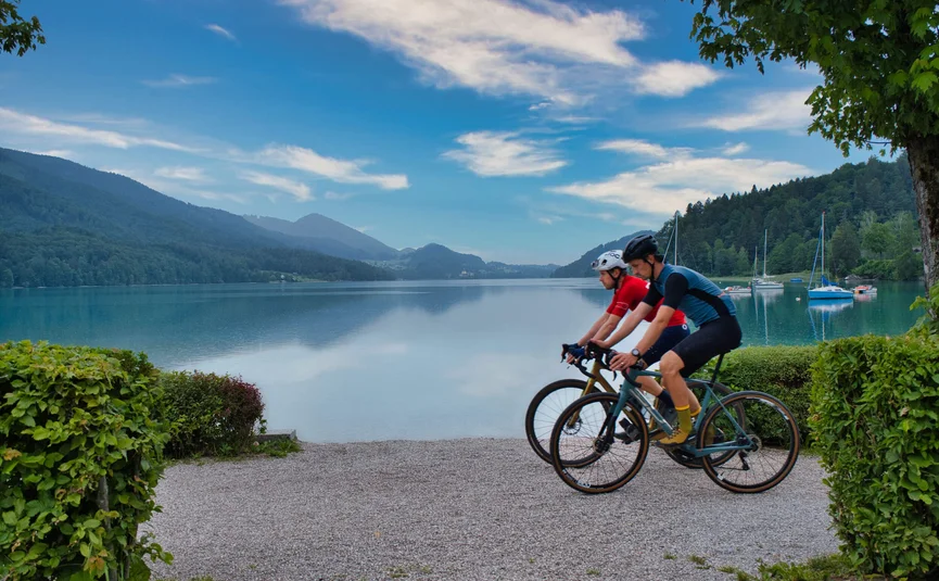 Gravelbike Urlaub in der Fuschlseeregion © Fuschlsee Tourismus - GravGrav Zwei Radfahrer fahren am Seeufer vor Bergen und Booten bei blauem Himmel