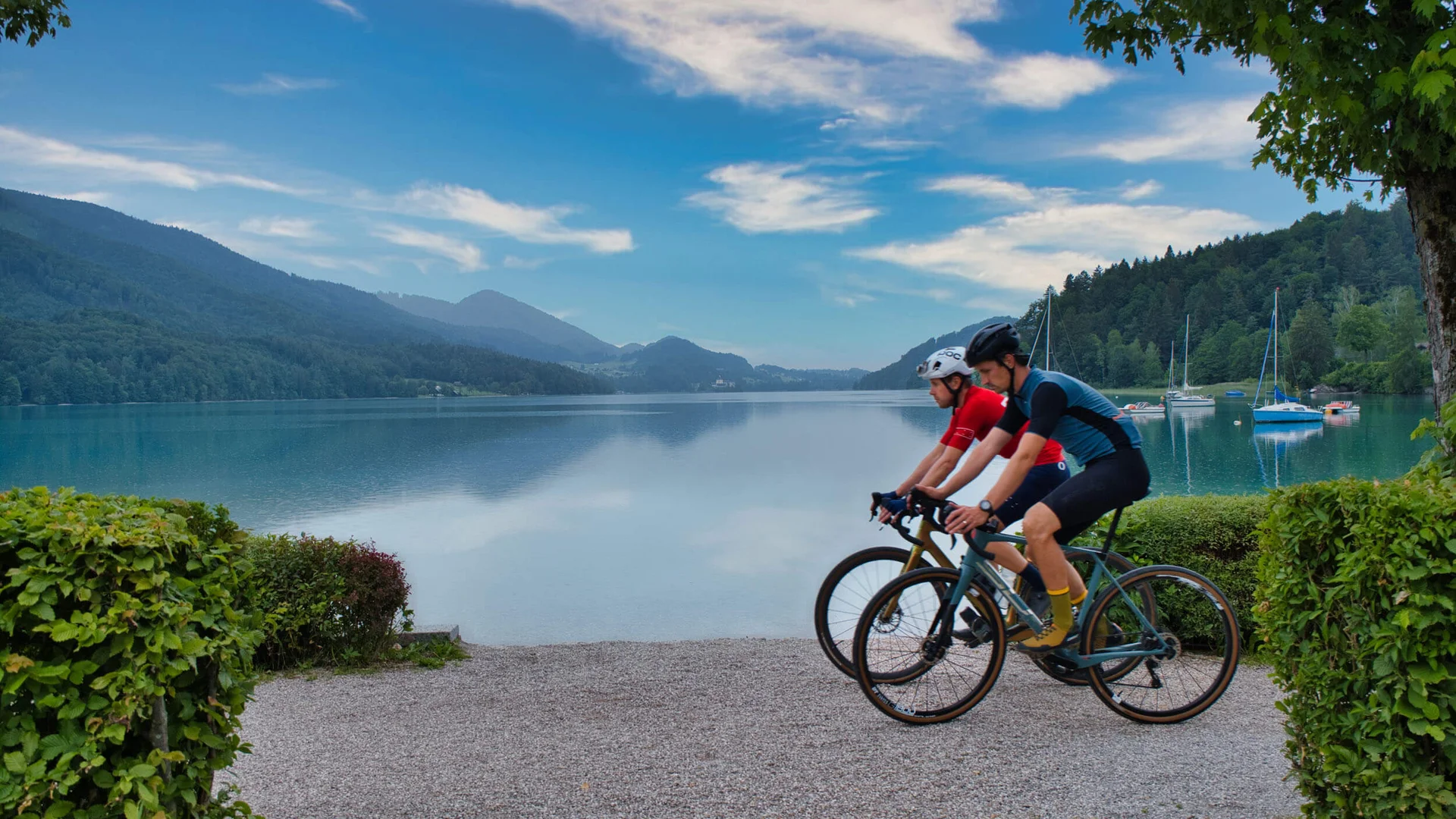 Zwei Radfahrer fahren am Seeufer vor Bergen und Booten bei blauem Himmel