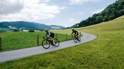 Two cyclists riding on a country road through green hills and forests.
