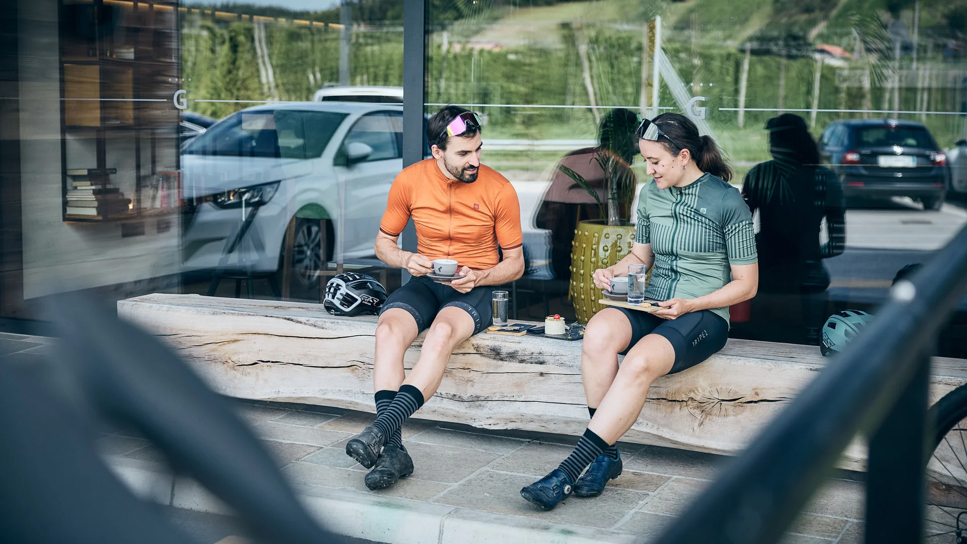 Zwei Radfahrer in Sportkleidung trinken Kaffee auf einer Holzbank draußen