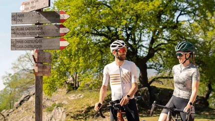Zwei Radfahrer sprechen an einem Wegweiser im Grünen bei Sonnenschein