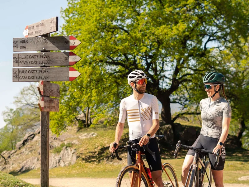 Gravelbike Holidays © Cinemepic / Teutschhaus Zwei Radfahrer sprechen an einem Wegweiser im Grünen bei Sonnenschein