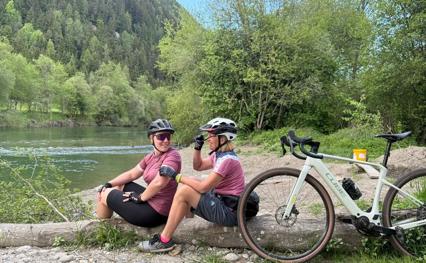 Zwei Radfahrerinnen machen am Flussufer eine Pause auf einem Baumstamm