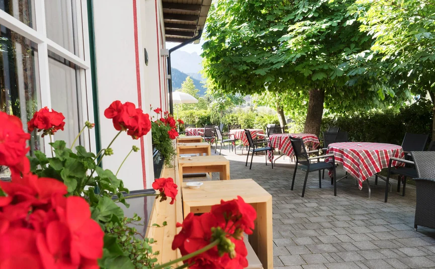 Terrasse mit roten Blumen, Tischen mit karierten Tischdecken und Stühlen unter Bäumen
