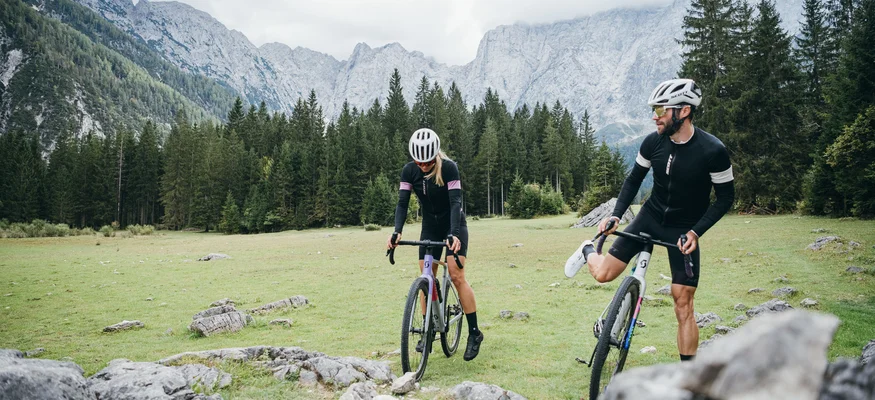 Zwei Radfahrer auf Mountainbikes in einer Bergwiese mit Nadelwald und Bergen