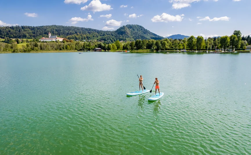 Zwei Personen stehend auf Paddleboards auf einem See mit Bergen im Hintergrund