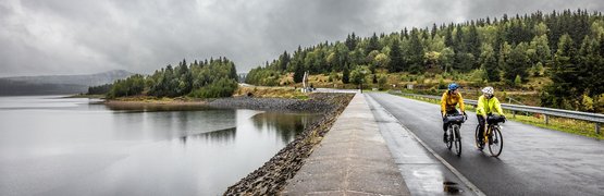 Two cyclists on wet road by lake and forested hill under cloudy sky