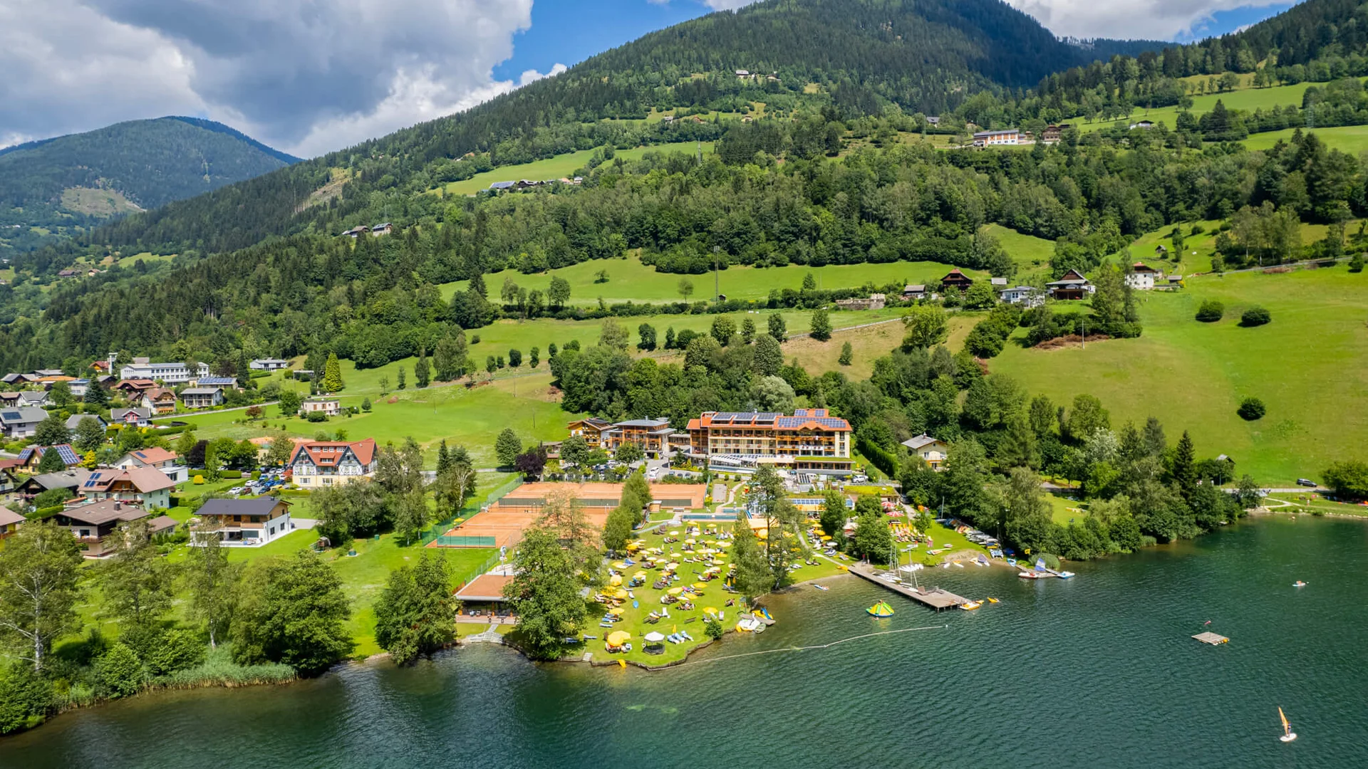 Lake shore with beach, sun loungers, and tennis courts by forested mountains