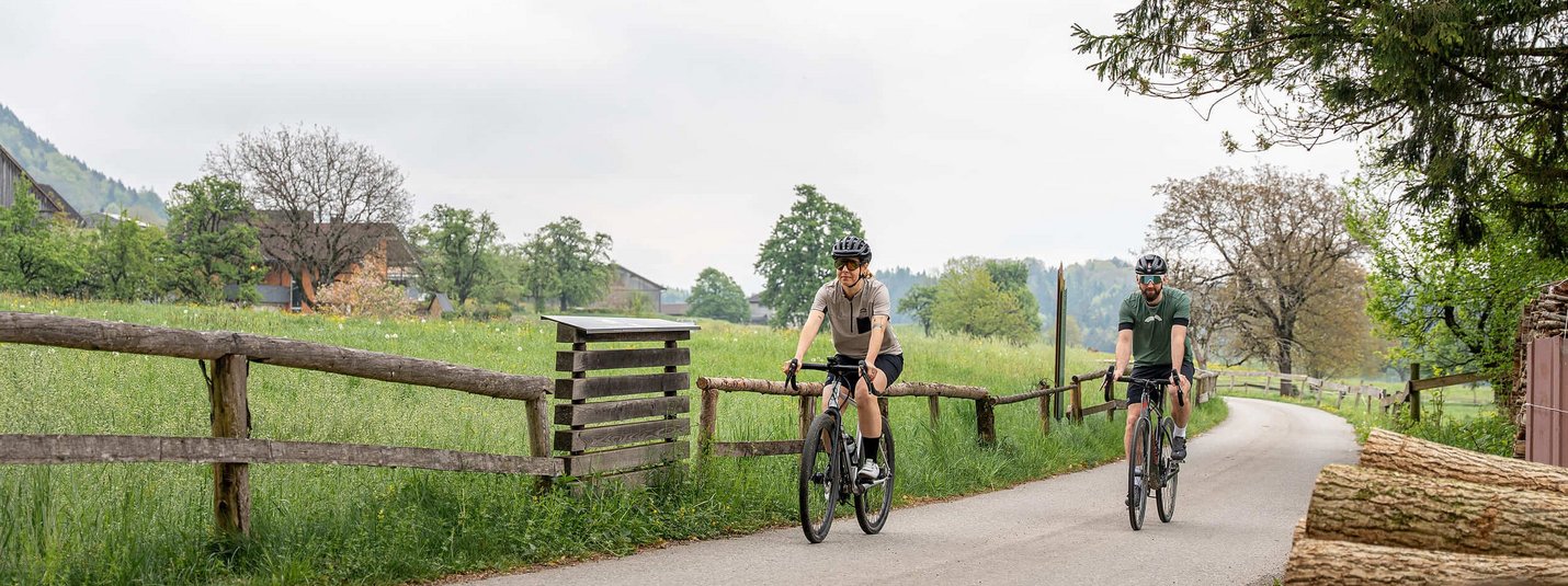 Bludenz Gravel Tour © Chris Gollhofer - Vorarlberg Tourismus Two cyclists on a country road with green fields and trees in the background