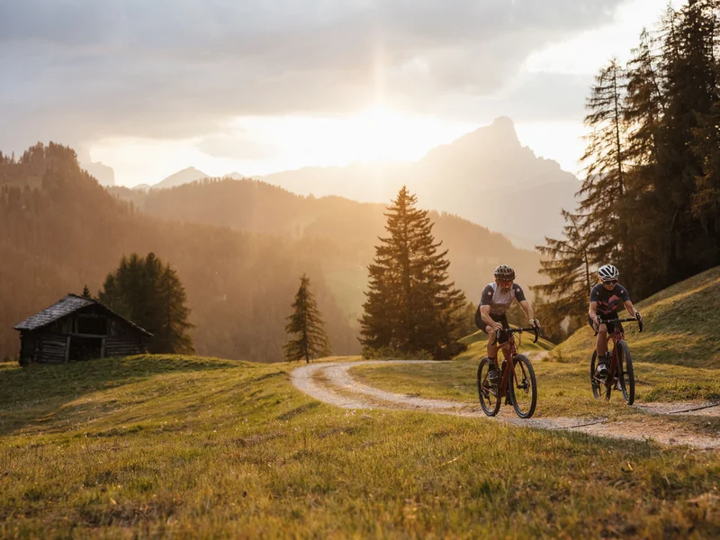 Gravelbike Holidays © Alex Moling Zwei Radfahrer fahren auf einem Waldweg bei Sonnenuntergang in den Bergen