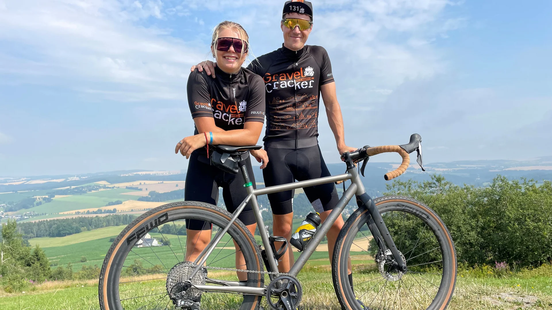 Zwei Radfahrer in Gravel Cracker Trikots mit silbernem Rennrad in Landschaft