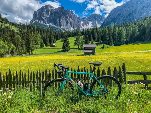 Gravel & Genuss am Kronplatz © Hotel Ostaria Posta Fahrrad an Holzzaun vor Blumenwiese und Berglandschaft bei sonnigem Himmel