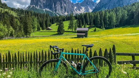 Gravel & Genuss am Kronplatz © Hotel Ostaria Posta Fahrrad an Holzzaun vor Blumenwiese und Berglandschaft bei sonnigem Himmel