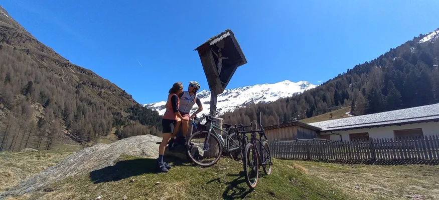 Radfahrer und Begleiter vor Bergkreuz mit schneebedeckten Alpen