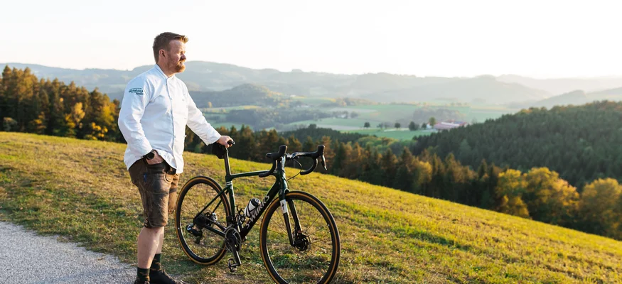 Am Krumbacherhof biked der Wirt! © Niederösterreich Werbung - Mara Hohla Mann steht mit Rennrad auf Landstraße mit Blick auf Hügel und Wald