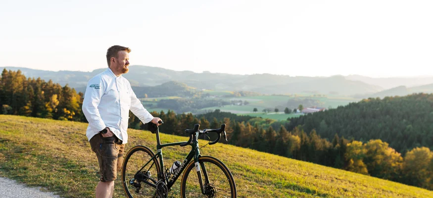 Mann steht mit Rennrad auf Landstraße mit Blick auf Hügel und Wald