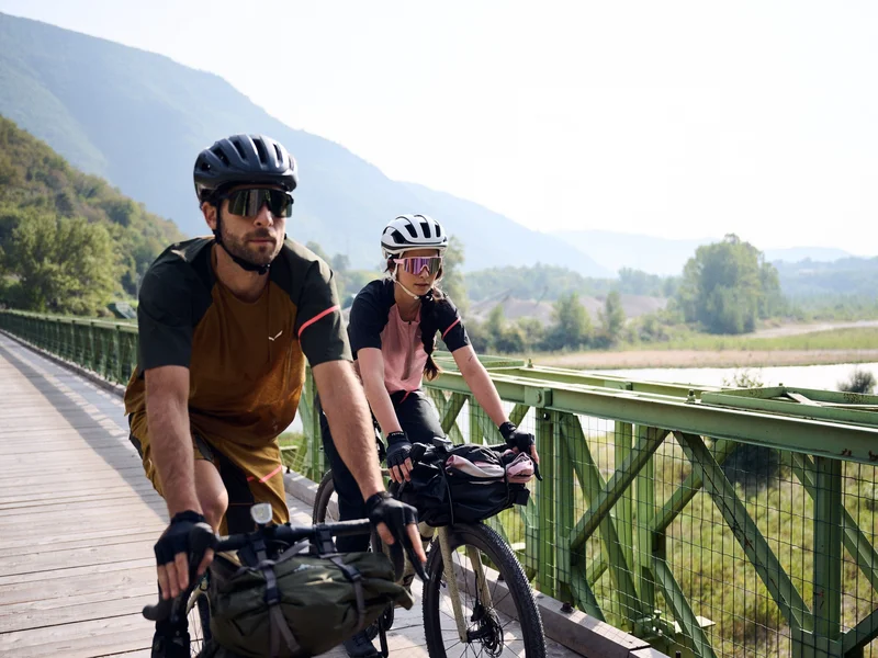 Zwei Radfahrer auf einer Brücke vor bewaldeten Hügeln und Flusslandschaft