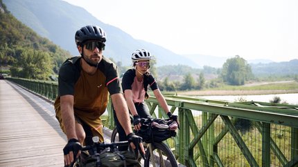 Zwei Radfahrer auf einer Brücke vor bewaldeten Hügeln und Flusslandschaft