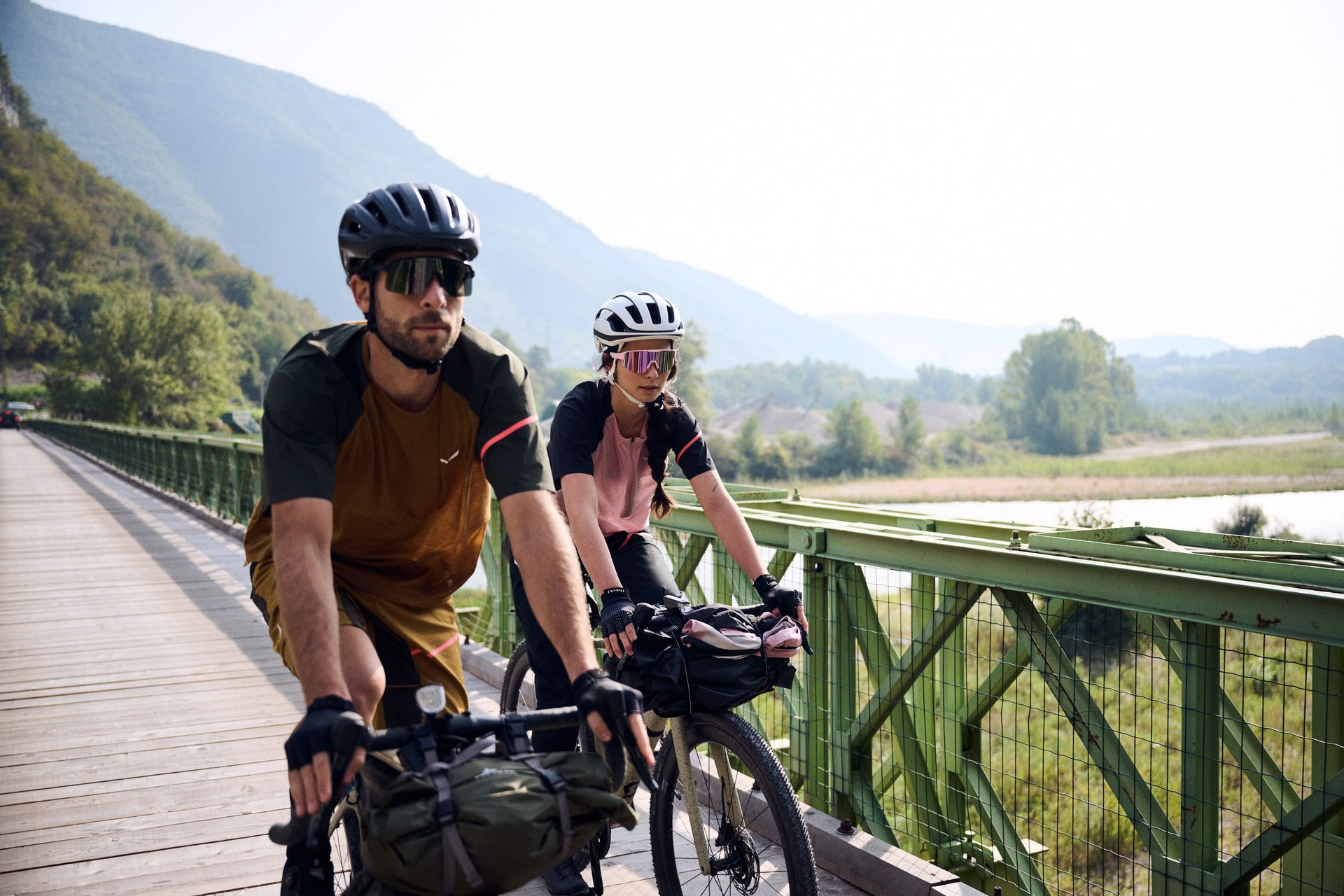 Zwei Radfahrer auf einer Brücke vor bewaldeten Hügeln und Flusslandschaft