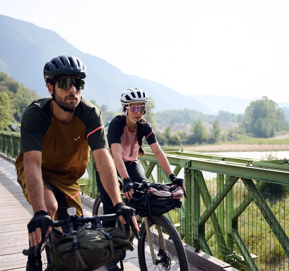 Zwei Radfahrer auf einer Brücke vor bewaldeten Hügeln und Flusslandschaft