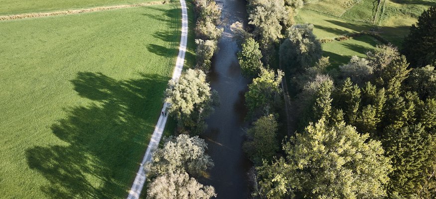 Luftaufnahme eines Flusses mit Bäumen und einem Weg daneben in einer ländlichen Umgebung