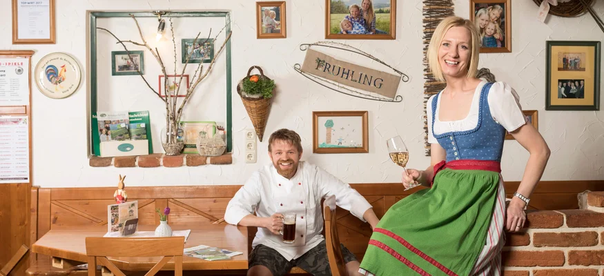 Am Krumbacherhof biked der Wirt! © Kurt Pinter Lächelnder Mann mit Bier und Frau im Dirndl mit Wein in rustikalem Gastraum