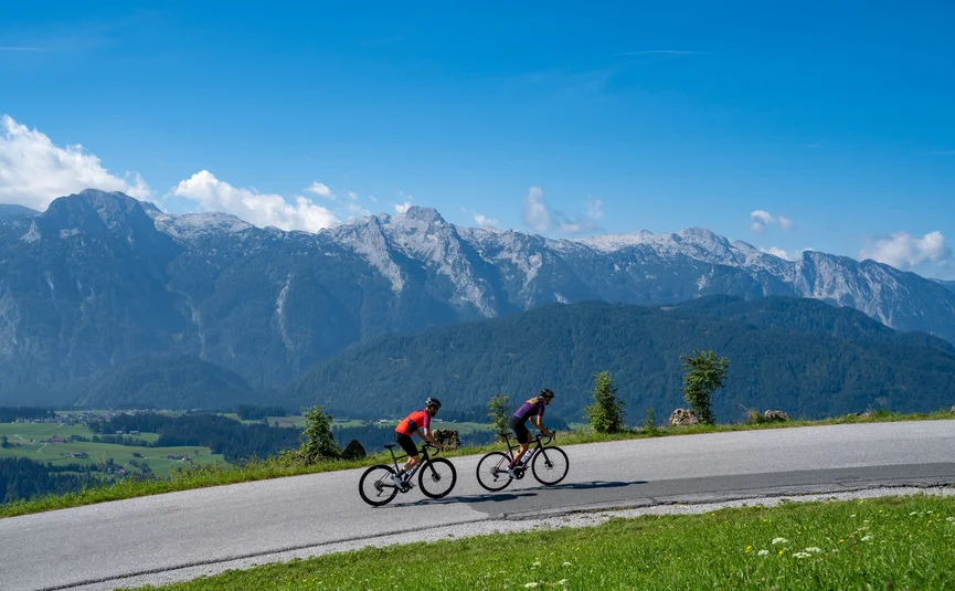 Gravelbike Urlaub in der Fuschlseeregion © Christina Neubauer Zwei Radfahrer auf Bergstraße vor Alpenpanorama bei klarem Himmel
