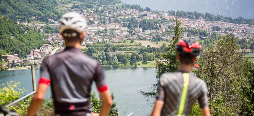 Zwei Radfahrer blicken auf einen See und eine Stadt in den Bergen