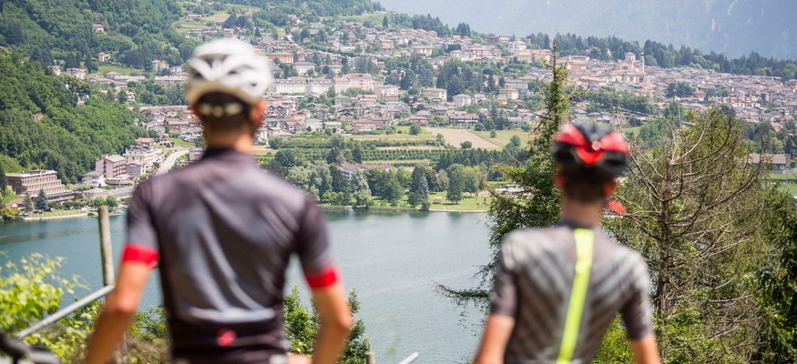 Zwei Radfahrer blicken auf einen See und eine Stadt in den Bergen