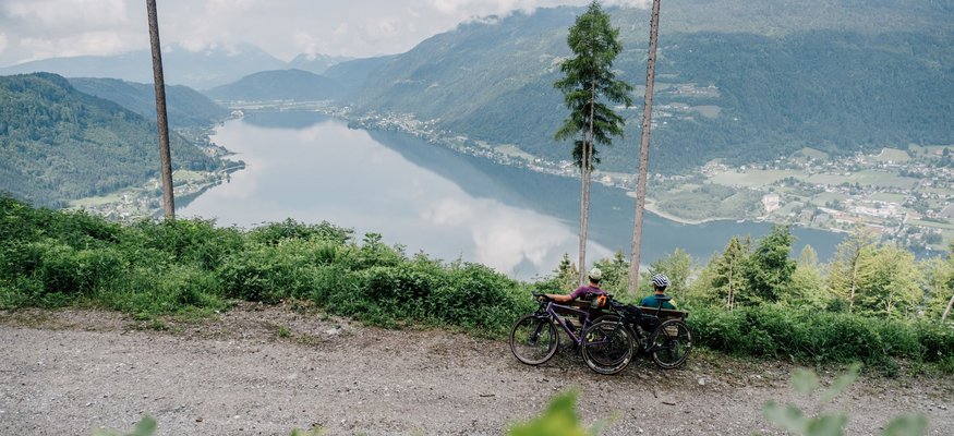 Zwei Radfahrer sitzen auf einem Bergweg mit Blick auf einen See und Berge.