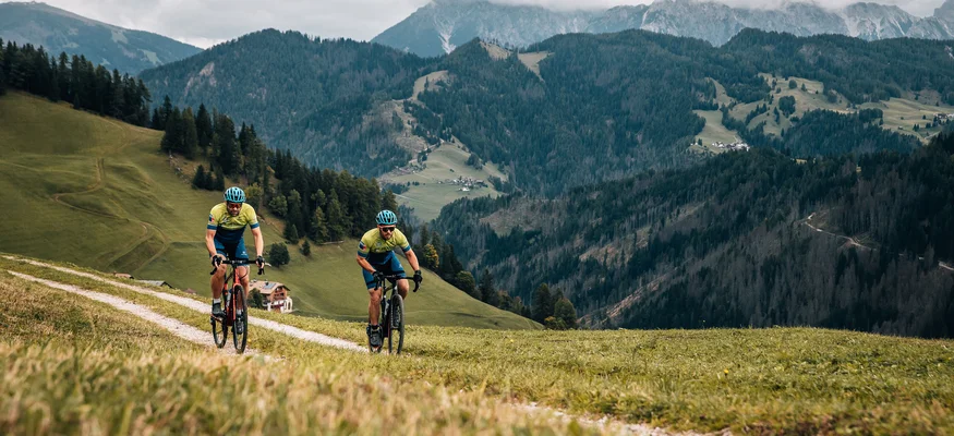 Zwei Radfahrer fahren auf einem Bergweg mit Wald und Bergen im Hintergrund