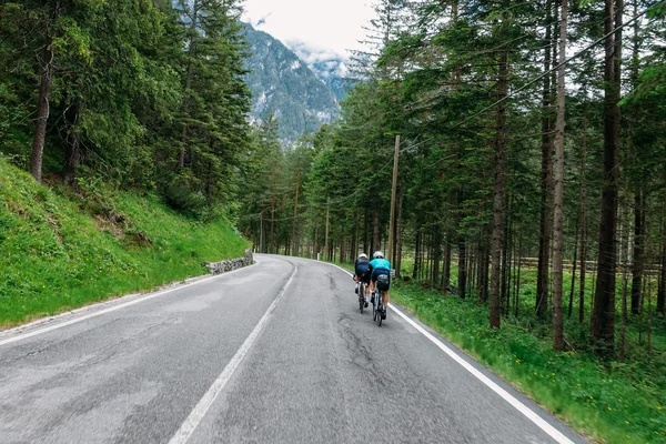 5 Gravelbike-Touren in Alta Badia, die du gefahren haben solltest © Hotel Melodia del Bosco Zwei Radfahrer auf bergiger Straße durch einen Wald mit Bergen im Hintergrund