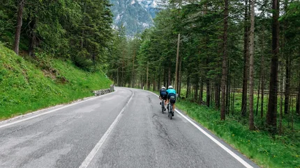 Zwei Radfahrer auf bergiger Straße durch einen Wald mit Bergen im Hintergrund