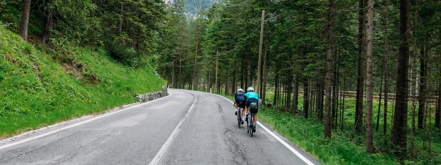 Austria Giro unsupported © Hotel Melodia del Bosco Zwei Radfahrer auf bergiger Straße durch einen Wald mit Bergen im Hintergrund