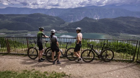Gravel & Törggelen: Warum Tramin im Herbst perfekt für Gravelbiker ist © Heiko Mandl Drei Radfahrer stehen mit ihren Fahrrädern an einem Geländer und blicken auf einen See und Berge.