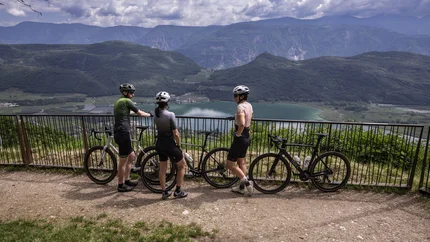 Three cyclists with bikes at a railing overlooking a lake and mountains.
