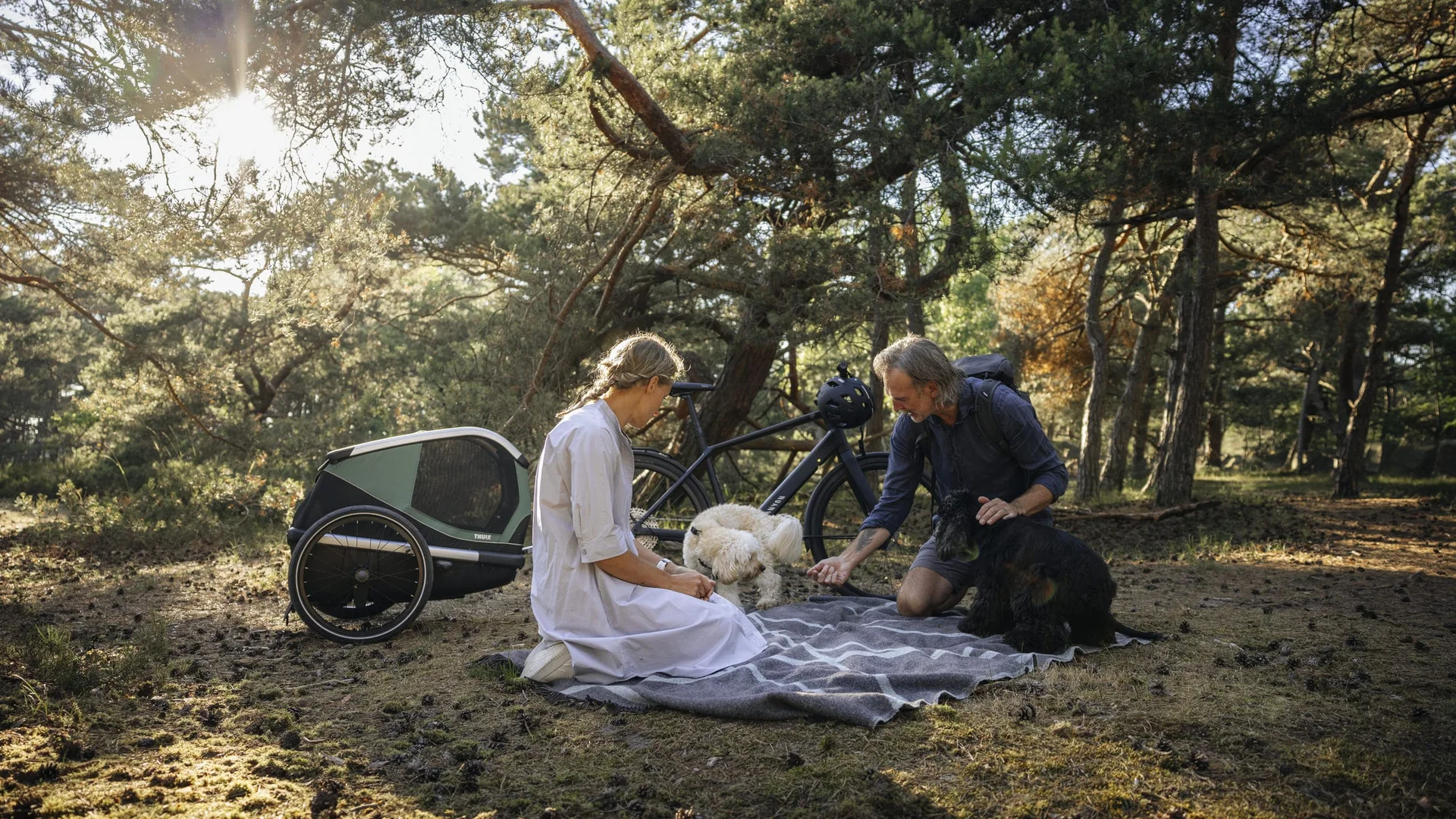 Mit dem Thule Bexey beginnt jedes Abenteuer zu zweit © Thule Zwei Personen sitzen mit zwei Hunden auf einer Decke im Wald neben einem Fahrradanhänger