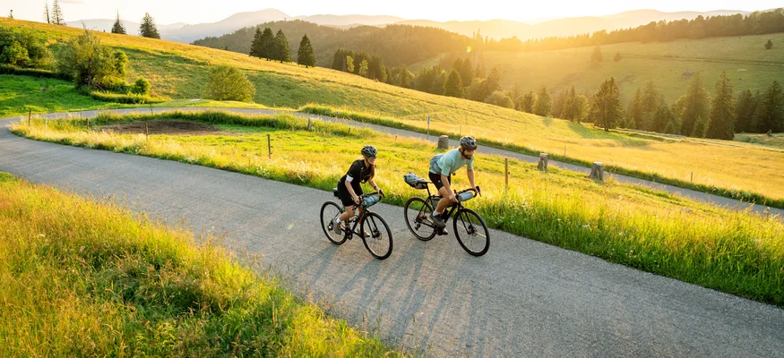 Posthorn Hotel-Restaurant © Hochschwarzwald Tourismus GmbH - Baschi Bender Zwei Radfahrer fahren bei Sonnenuntergang auf einer Straße durch grüne Hügel