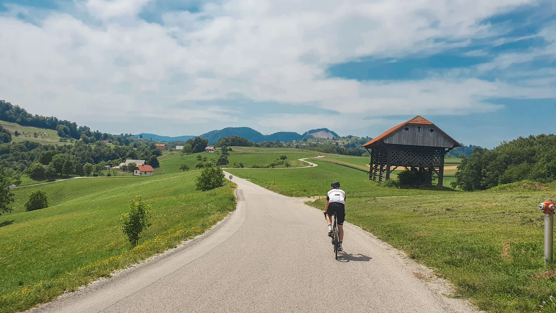 Radfahrer auf Landstraße mit grünem Hügel und traditionellem Holzbau im Hintergrund