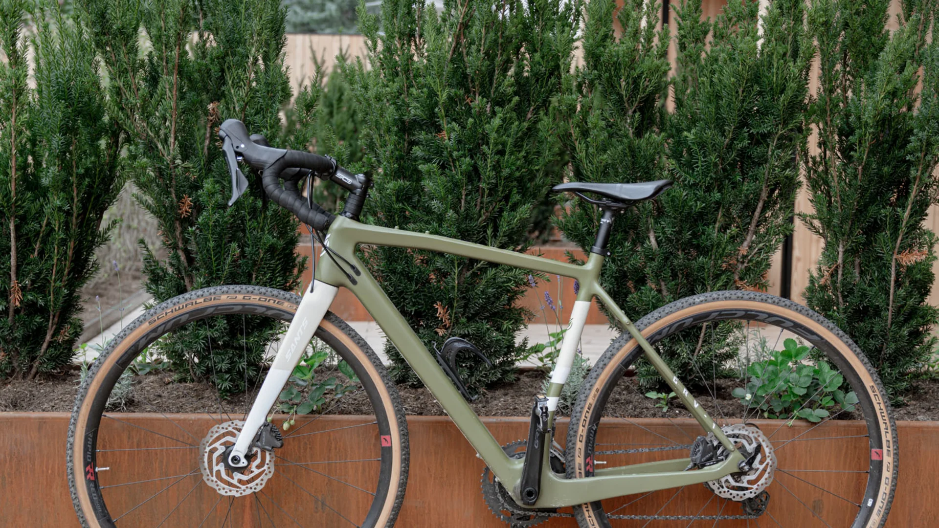 Grünes Gravelbike lehnt an rustikalem Pflanzkasten vor moderner Holzfassade