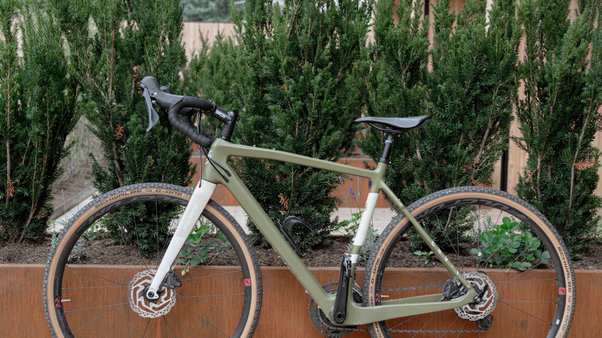 Grünes Gravelbike lehnt an rustikalem Pflanzkasten vor moderner Holzfassade