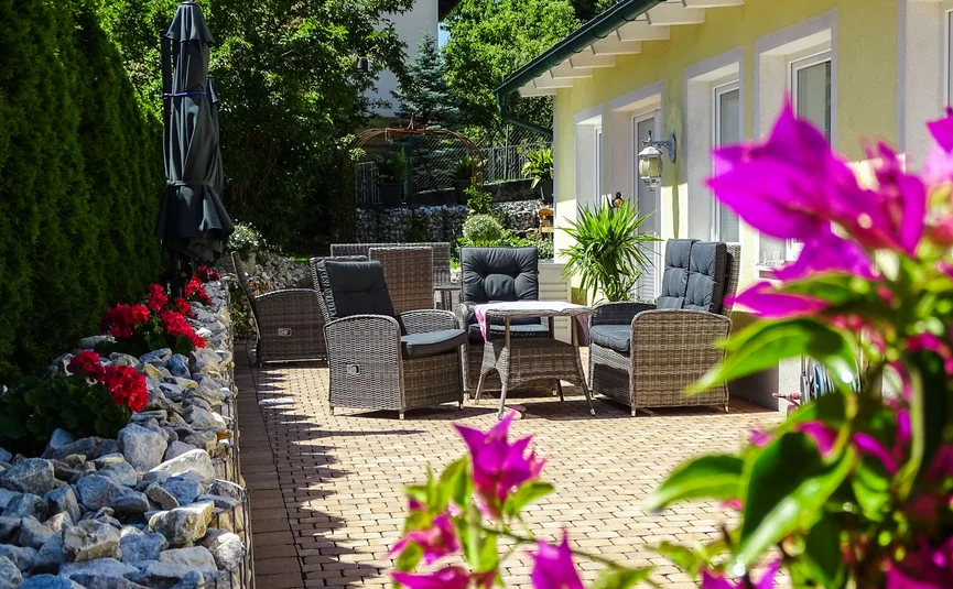 Sitzbereich mit Rattanstühlen neben gelbem Haus und blühenden Pflanzen