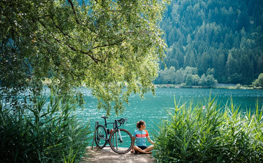 Bike Boutique Hotel Lindenhof **** © Martin Hofmann Frau sitzt am Seeufer neben Fahrrad unter grünem Baum im Sommer