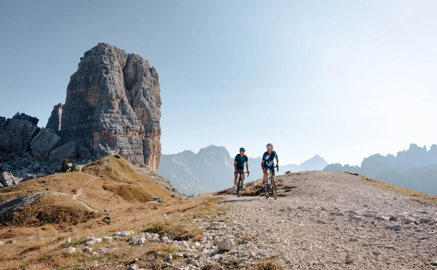 Two cyclists on mountain trail with large rock formation and mountain backdrop