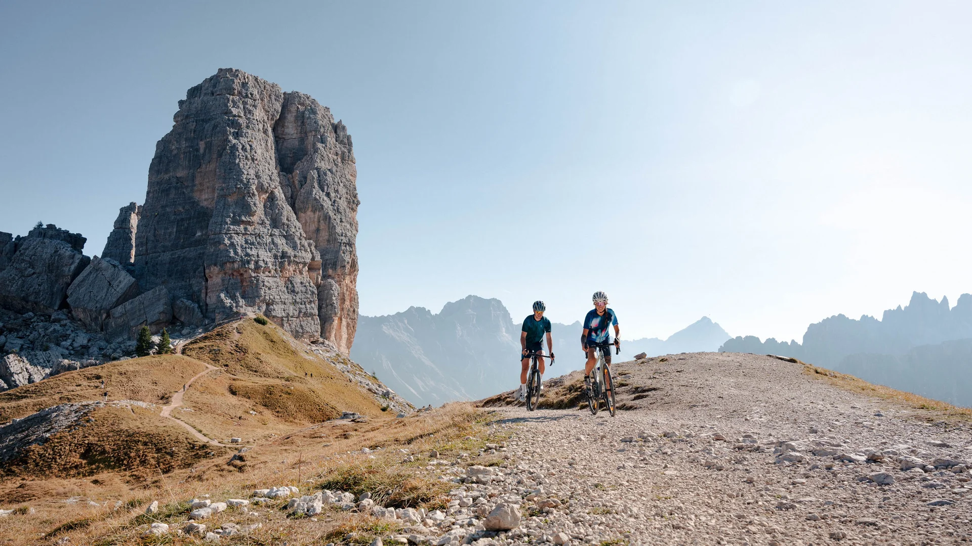 Zwei Radfahrer auf Bergweg mit großem Felsen und Bergkulisse im Hintergrund