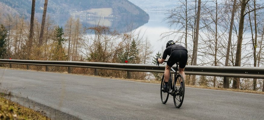 Hotel zur Post ***S © Martin Hoffmann Cyclist riding downhill on paved road with lake and mountains in background