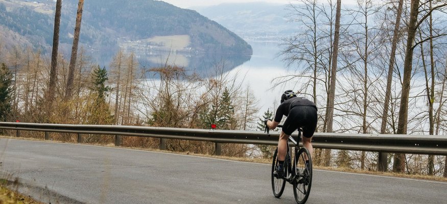 Radfahrer fährt bergab auf asphaltierter Straße mit See und Bergen im Hintergrund