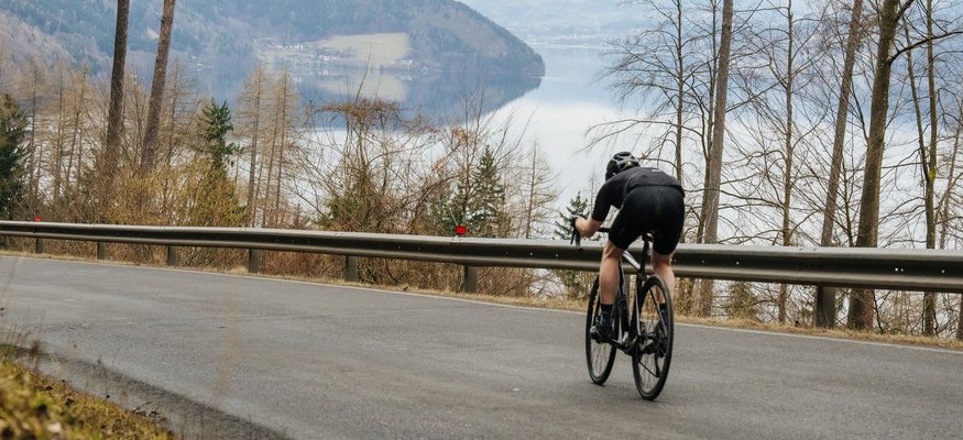 Radfahrer fährt bergab auf asphaltierter Straße mit See und Bergen im Hintergrund